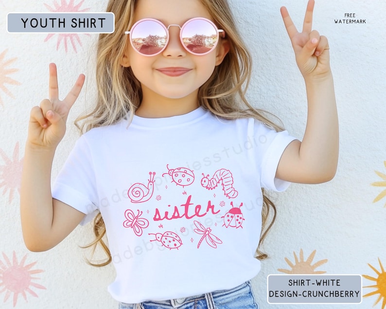 Op de afbeelding: Wit kinder-T-shirt met een roze afbeelding van insecten en het woord "sister". De persoon die het T-shirt draagt, draagt een roze zonnebril en maakt een vredesteken met haar vingers.