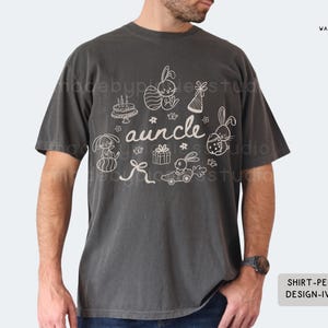 May include: Dark grey t-shirt with a white graphic featuring the word "auncle" and Easter-themed illustrations, including bunnies, eggs, and a cake. The shirt has a relaxed fit and short sleeves.
