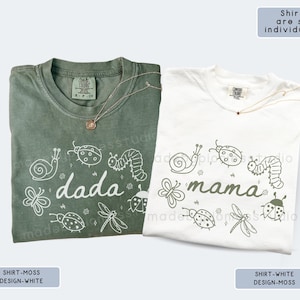 Op de afbeelding: Twee T-shirts, &eacute;&eacute;n mosgroen en &eacute;&eacute;n wit, elk met een uniek ontwerp. Het mosgroene shirt heeft het woord "dada" met insectenillustraties. Het witte shirt toont "mama" met vergelijkbare insectengraphics. Beide shirts hebben een ronde hals.