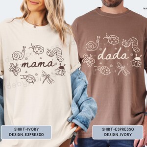 Op de afbeelding: Twee T-shirts, &eacute;&eacute;n ivoor en &eacute;&eacute;n espresso, elk met een uniek ontwerp. Het ivoorkleurige shirt toont het woord "mama" omringd door insectenillustraties. Het espresso-shirt toont het woord "dada" met vergelijkbare insectenkunst. Beide shirts zijn vrijetijdskleding.