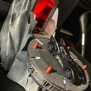 Può includere: Un'auto giocattolo nera con un simbolo di pipistrello sul cofano è su una piattaforma grigia con coni rossi. La piattaforma fa parte di una struttura grigia più grande che assomiglia a una montagna.