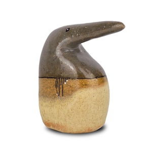Può includere: Una statuetta di uccello in ceramica con un becco lungo e sottile e una glassa marrone e beige. L'uccello è in piedi su uno sfondo bianco.