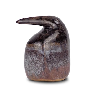 May include: A handcrafted, ceramic sculpture in the shape of a bird. The sculpture has a dark brown, glossy finish on the head and beak, transitioning to a textured, purplish-brown body. The beak extends horizontally from the head.
