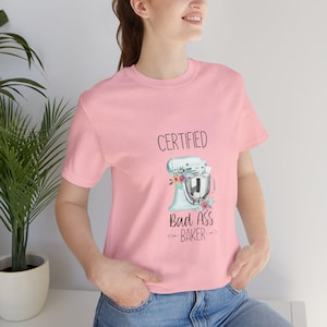 May include: A light pink t-shirt featuring the text "CERTIFIED Bad Ass BAKER" above a light blue stand mixer design with floral accents. The shirt is made of soft material and is suitable for casual wear.