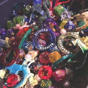 May include: A collection of colorful dice, rings, and small figurines. The dice are various shapes and colors, including red, green, blue, and yellow. Some rings have text that reads "BLINDED" and "DEAFENED". A blue dragon figurine is also visible.