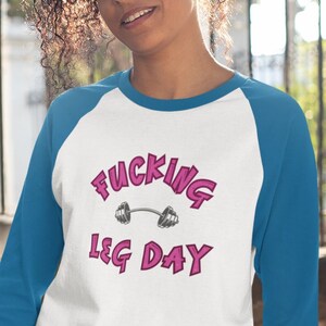May include: A white and blue baseball tee shirt with the text "FUCKING LEG DAY" in pink lettering and a pink barbell graphic.