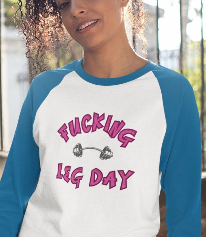 May include: A white and blue baseball tee shirt with the text "FUCKING LEG DAY" in pink lettering and a pink barbell graphic.