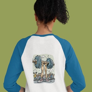 May include: A white and blue baseball tee shirt with a cartoon illustration of a person lifting a barbell with a crowd of people cheering in the background.