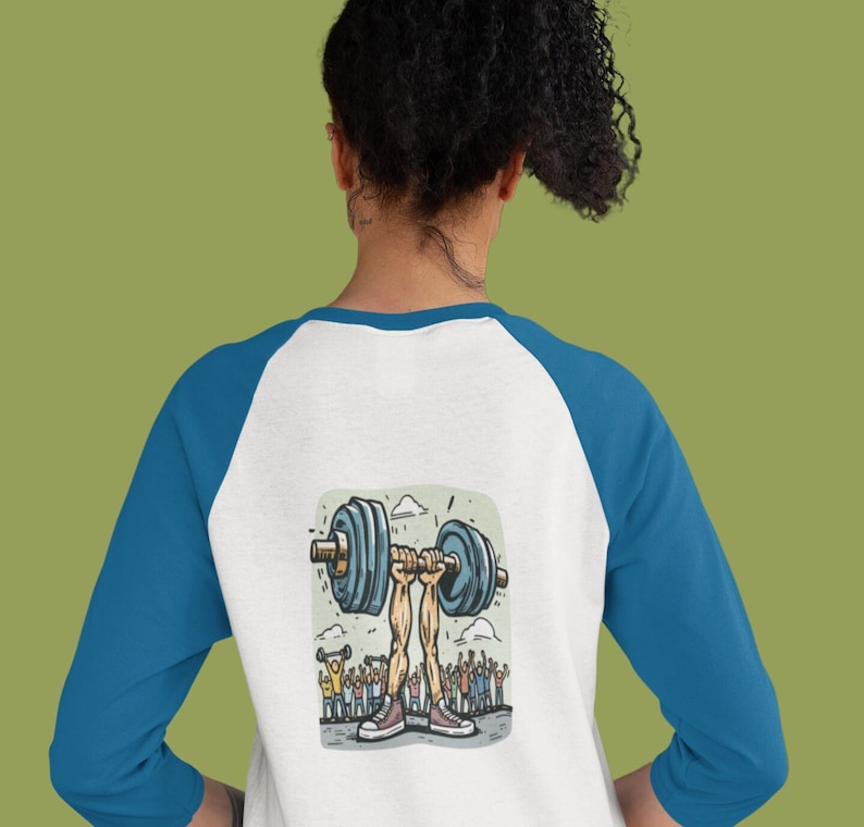 May include: A white and blue baseball tee shirt with a cartoon illustration of a person lifting a barbell with a crowd of people cheering in the background.