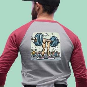 May include: A grey and red baseball t-shirt featuring a cartoon illustration of a pair of legs lifting a barbell with a crowd of people cheering in the background.