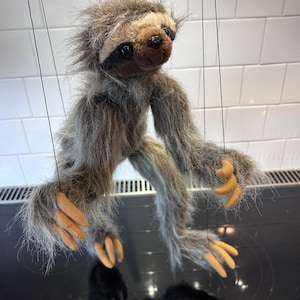 May include: A sloth marionette puppet featuring long, shaggy grey and brown faux fur, a dark brown face mask, and black eyes. The puppet has tan, claw-like hands and feet, and is suspended by thin black strings attached to its limbs and head.