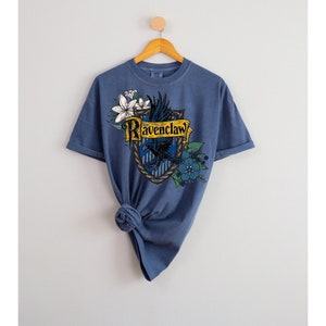 Könnte beinhalten: Ein blaues T-Shirt mit einem Aufdruck des Wappens des Ravenclaw-Hauses aus der Harry-Potter-Reihe. Das Wappen zeigt einen blauen und goldenen Schild mit einem Raben und den Worten "Ravenclaw" in Gold.