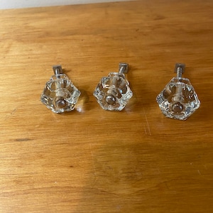 Puede incluir: Tres pomos de armario de cristal transparente con bases y tornillos de metal plateado. Los pomos tienen un diseño facetado.