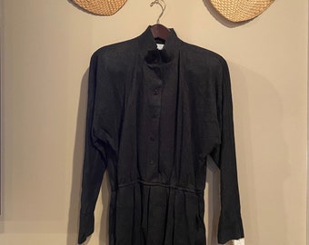 May include: A dark gray dress with long sleeves and a button-up front hangs on a wooden hanger. The dress has a high neckline and a gathered waist. Two woven fans are mounted on the wall behind the dress.