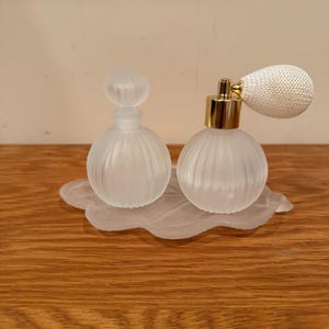 Puede incluir: Dos botellas de perfume de vidrio esmerilado con tapas doradas y una boquilla de pulverización blanca sobre una bandeja de vidrio esmerilado en forma de hoja.