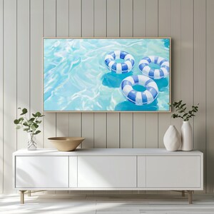 May include: A framed artwork depicts three blue and white striped inflatable rings floating in a pool of light blue water. The painting is displayed above a white cabinet with a wooden bowl and decorative vases.