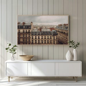 May include: A framed print of a European cityscape, featuring buildings with gray rooftops and red chimneys under a cloudy sky. The artwork hangs above a white cabinet with decorative vases and a bowl.