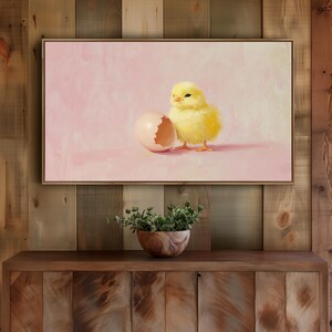May include: A framed painting depicts a fluffy yellow chick standing next to a cracked eggshell on a pink background. The artwork has a soft, painterly style, with visible brushstrokes. The painting is displayed above a wooden cabinet.