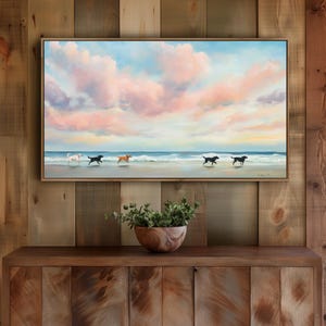 Puede incluir: Una pintura enmarcada representa perros corriendo en una playa al atardecer. El cielo está lleno de nubes rosas y azules. Los perros son de varios colores, incluyendo blanco, negro y marrón. La pintura se exhibe sobre un mueble de madera.