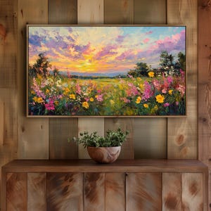 May include: A framed landscape painting shows a sunset over a field of wildflowers. The sky has pink, yellow, and blue colours, and the foreground features colourful blooms. The painting is displayed on a wooden cabinet.