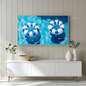 Puede incluir: Una pintura enmarcada representa dos flotadores inflables a rayas azules y blancas flotando en una piscina. El agua es de un azul vibrante con reflejos de luz. La obra de arte se exhibe sobre un mueble blanco.