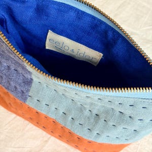 May include: A zipped pouch featuring a patchwork design of orange, light blue, and dark grey fabric panels with visible dark blue running stitch embroidery. The interior is lined with bright blue fabric and includes a white label reading eelo & ider.