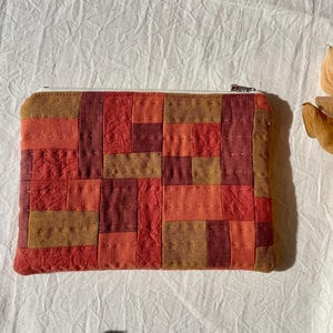 May include: A rectangular zipped pouch featuring a patchwork design of fabric rectangles in shades of rust, burnt orange, mustard yellow, and deep red. The surface is accented with visible, straight-line hand-stitched quilting details.