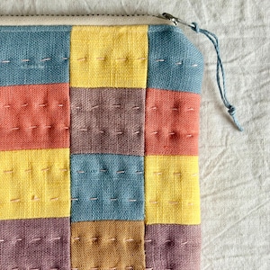 May include: A zipped pouch featuring a patchwork design of square fabric panels in shades of blue, yellow, terracotta, and muted purple. The textile surface is accented with visible light-coloured running stitches throughout, and a blue string pull.