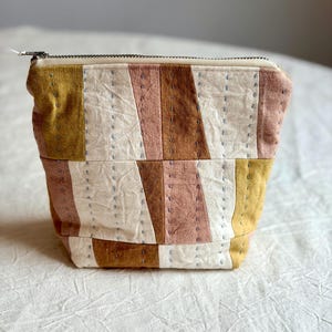 May include: A zipped pouch featuring a patchwork design of mustard yellow, dusty pink, brown, and off-white fabric panels. The exterior is decorated with vertical rows of light blue running stitches that create a textured, quilted appearance.
