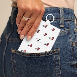May include: A white keychain with a red cherry pattern and the letter "S" in the center. The keychain is being held in a back pocket of blue jeans.