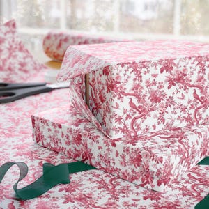 May include: Two gift boxes wrapped in white paper with a red floral pattern. The boxes are stacked on top of each other and are tied with a green ribbon.