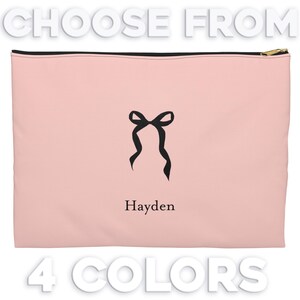 May include: A pink zippered pouch with a black bow and the name Hayden printed on it. The pouch is available in four colors.