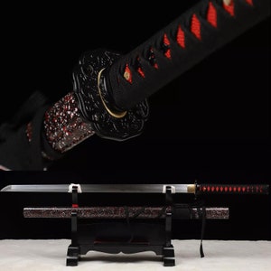 May include: A Japanese katana sword with a black handle wrapped in red diamond-patterned fabric. The blade is silver, and the scabbard is black with red speckles. The sword rests on a black stand against a dark background.