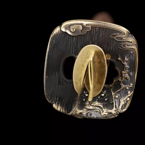 May include: Close-up of a Japanese katana tsuba, a hand guard. The tsuba is dark with gold accents, featuring a floral and cloud design. The central opening is for the sword's tang. The guard is made of metal.
