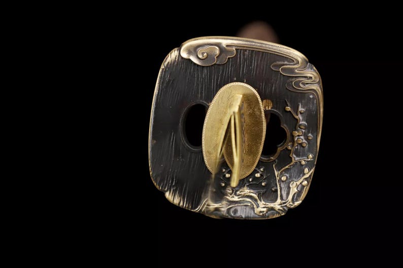 May include: Close-up of a Japanese katana tsuba, a hand guard. The tsuba is dark with gold accents, featuring a floral and cloud design. The central opening is for the sword's tang. The guard is made of metal.
