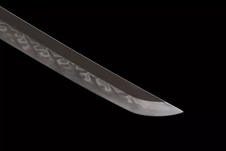 May include: Close-up of a katana blade with a polished, reflective surface. The blade has a subtle wave pattern along its length, and the tip is sharply pointed. The background is solid black, emphasizing the blade's form.