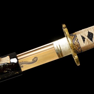 Pode incluir: Um close-up de uma espada katana dourada com uma bainha preta e dourada. A espada tem um desenho decorativo de um drag&atilde;o na l&acirc;mina. O cabo &eacute; envolto em um cord&atilde;o bege com padr&otilde;es pretos em forma de diamante.