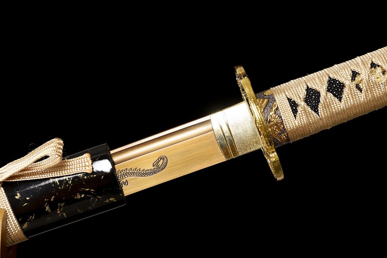 Pode incluir: Um close-up de uma espada katana dourada com uma bainha preta e dourada. A espada tem um desenho decorativo de um drag&atilde;o na l&acirc;mina. O cabo &eacute; envolto em um cord&atilde;o bege com padr&otilde;es pretos em forma de diamante.