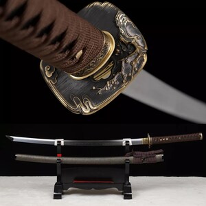 May include: An ornate katana sword with a black blade and a decorative handle. The handle is wrapped in brown fabric and features gold-colored accents. The sword is displayed on a black stand, with its scabbard visible below.