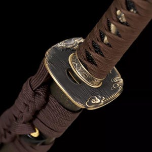 May include: Close-up of a Japanese katana sword hilt. The handle is wrapped in brown fabric, with a decorative black and gold guard. The guard features a cloud-like design. The sword's details are set against a black background.