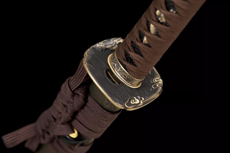 May include: Close-up of a Japanese katana sword hilt. The handle is wrapped in brown fabric, with a decorative black and gold guard. The guard features a cloud-like design. The sword's details are set against a black background.