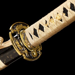 Pode incluir: Close-up de uma espada katana com uma empunhadura dourada e um envolt&oacute;rio tran&ccedil;ado branco e preto. A empunhadura &eacute; decorada com desenhos intrincados.