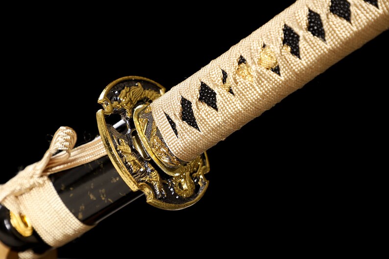 Pode incluir: Close-up de uma espada katana com uma empunhadura dourada e um envolt&oacute;rio tran&ccedil;ado branco e preto. A empunhadura &eacute; decorada com desenhos intrincados.