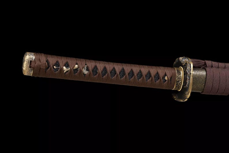 May include: Close-up of a katana handle, featuring a brown, tightly wrapped grip with diamond-shaped black accents. The handle's end has a gold-colored cap, and the guard is a dark metal with gold detailing. The background is solid black.