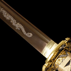 Pode incluir: Close-up de uma espada katana com uma empunhadura dourada e um desenho de drag&atilde;o gravado na l&acirc;mina.