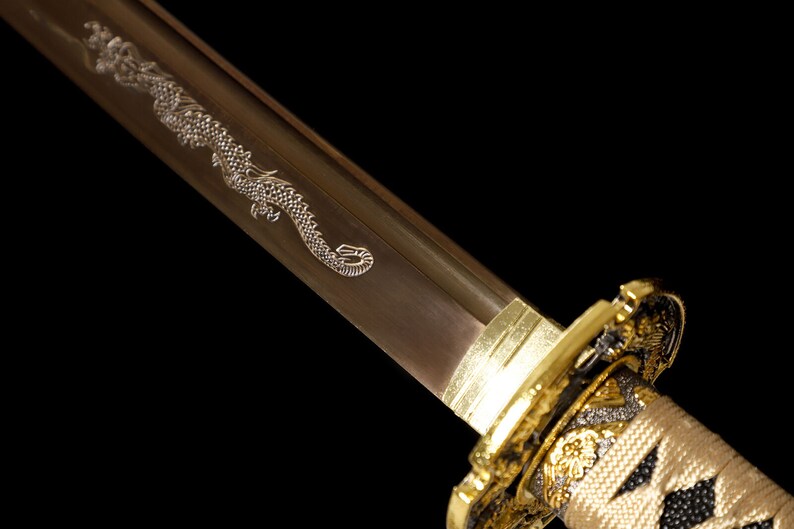 Pode incluir: Close-up de uma espada katana com uma empunhadura dourada e um desenho de drag&atilde;o gravado na l&acirc;mina.