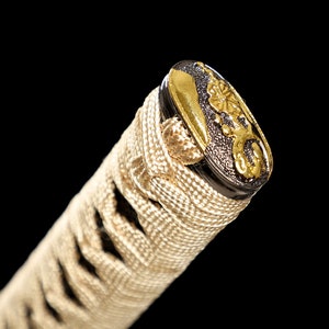 Pode incluir: Close-up da parte superior de um cabo de katana, ou tsuka, envolto em ito branco, um tipo de corda, com um menuki dourado e preto, um ornamento decorativo.