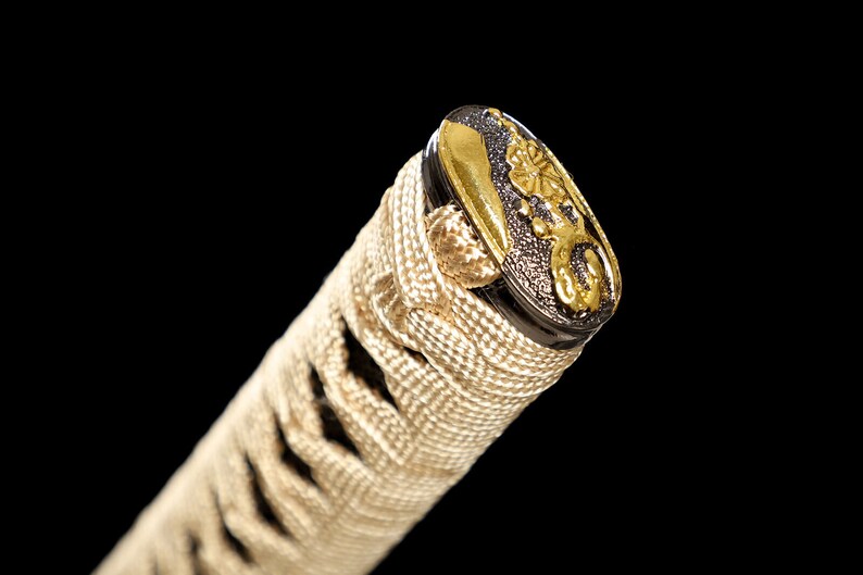 Pode incluir: Close-up da parte superior de um cabo de katana, ou tsuka, envolto em ito branco, um tipo de corda, com um menuki dourado e preto, um ornamento decorativo.