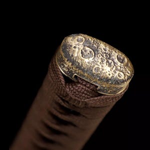 May include: Close-up of a brown handle with a textured, gold-colored cap. The cap has a pattern of small, irregular circles. The handle appears to be wrapped in a woven, brown material, suggesting a decorative or functional item.