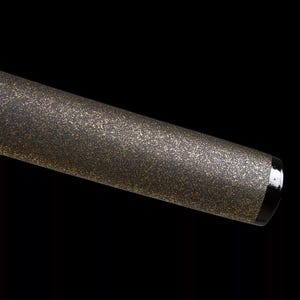 May include: Close-up of a metallic, cylindrical object with a speckled, textured surface in shades of gold and black. The object has a black band and a silver end cap. The background is solid black.